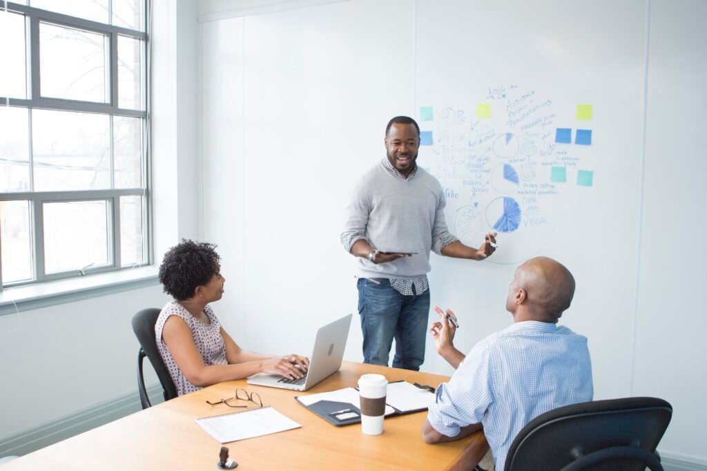 Three online marketing professionals having a collaborative meeting in a bright conference room. A man stands near a whiteboard, holding a tablet and explaining a concept, while a woman and another man sit at the table with a laptop and notes, actively engaging in the discussion. The whiteboard is filled with diagrams and sticky notes, and large windows flood the room with natural light, creating an inviting and productive environment.